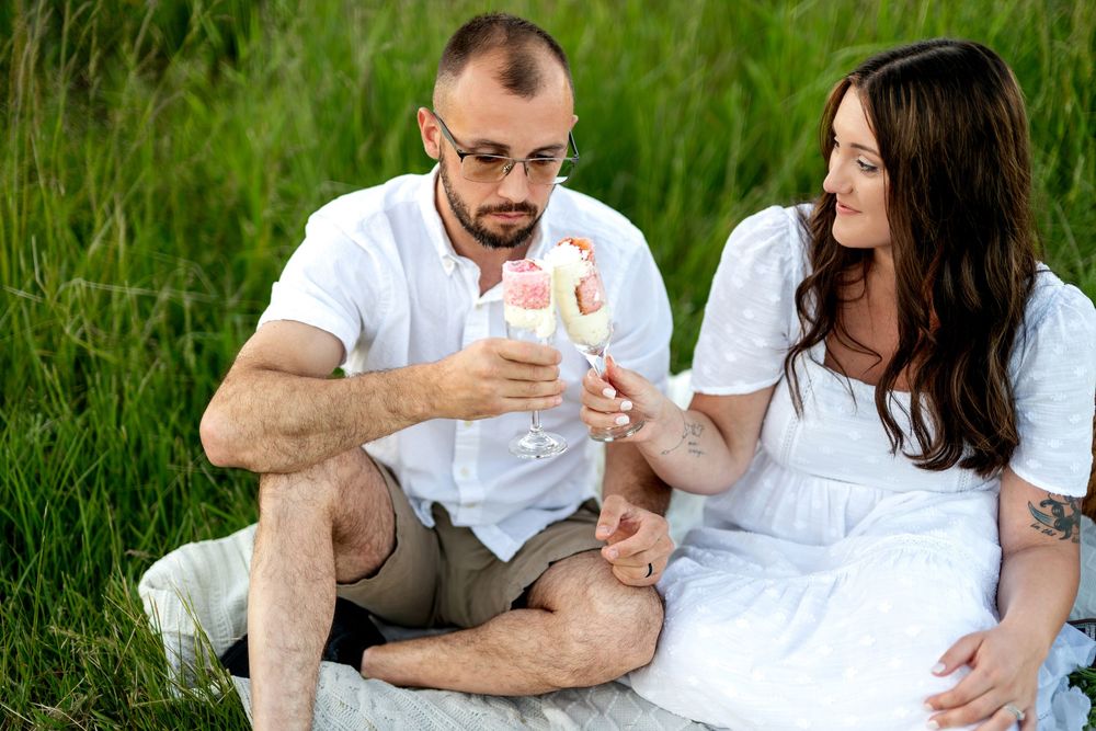 Couple sitting in green grass sharing champagne flutes during a romantic outdoor picnic engagement session.