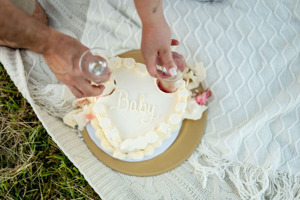 Couple cutting a white baby shower cake with floral decorations on a picnic blanket outdoors.