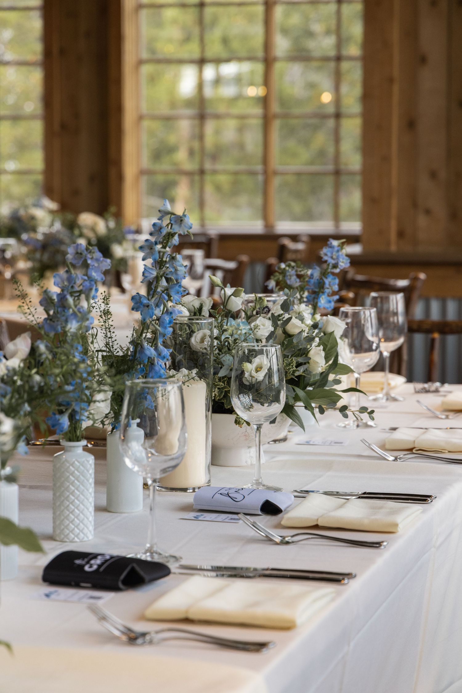 Elegant wedding reception table with blue floral centerpieces, white linens, crystal glassware, and gold accents in a rustic venue.
