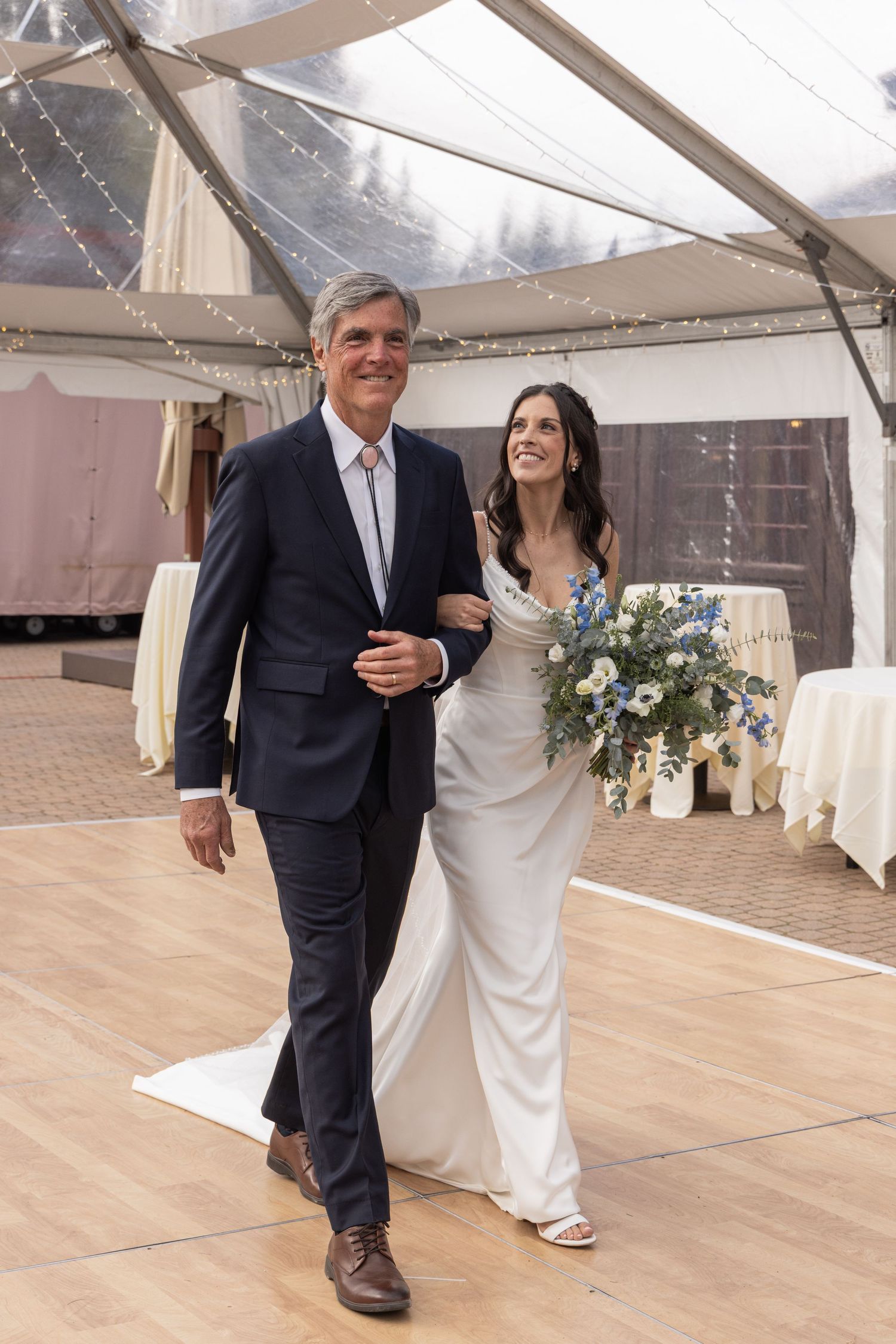 Bride in white gown holding blue floral bouquet walks arm-in-arm with suited man under elegant tent venue.