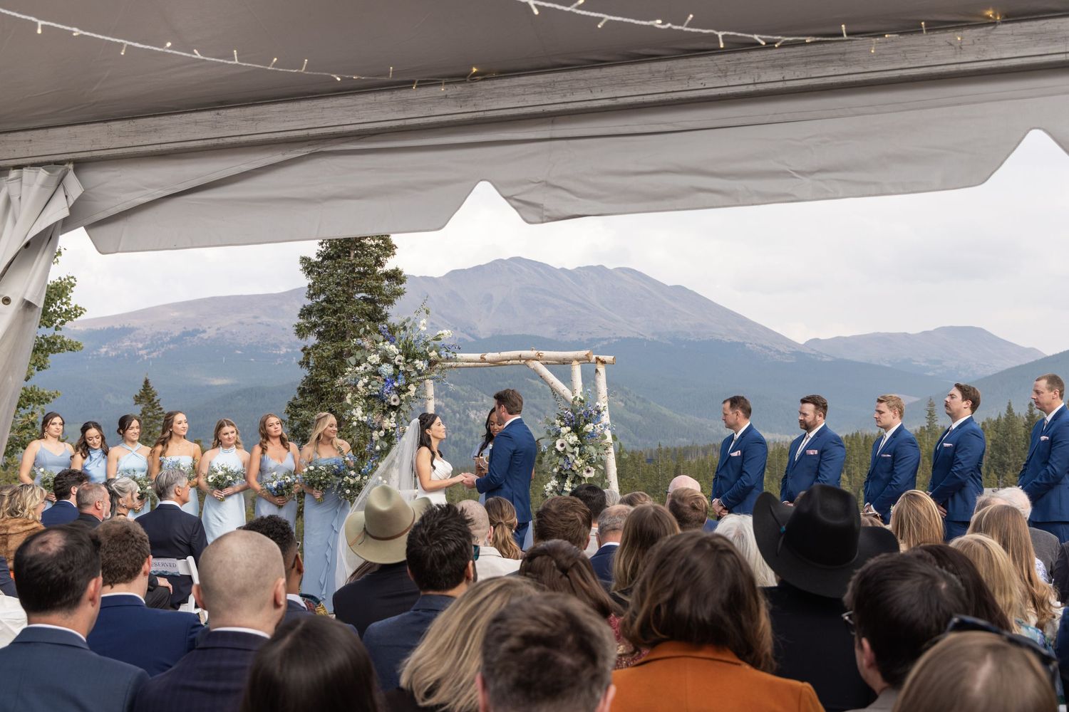 Outdoor mountain wedding ceremony under a white tent with bridal party in blue, stunning Rocky Mountain backdrop.