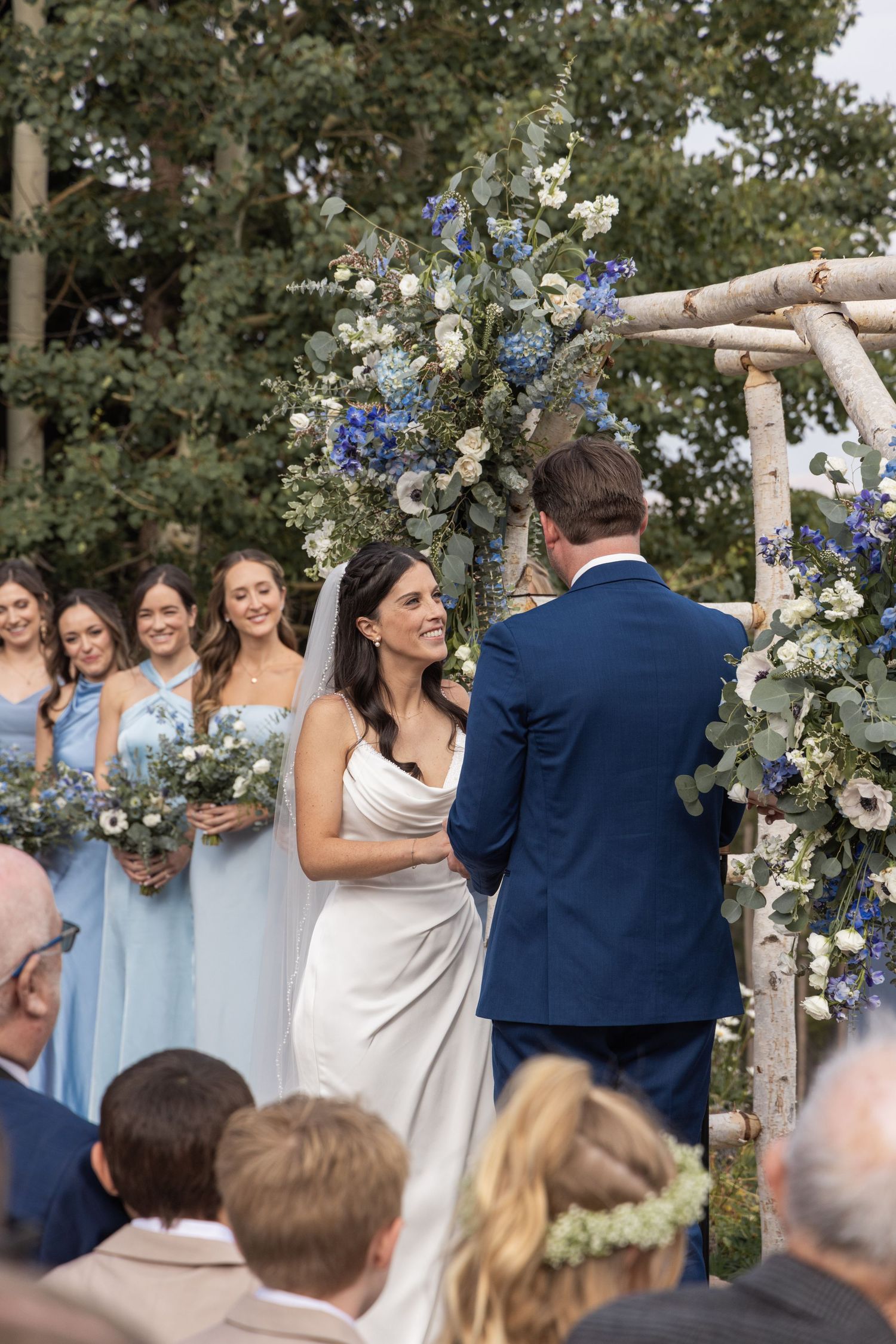 Bride in white gown smiles at groom during outdoor wedding ceremony beneath floral arch with bridesmaids in blue.