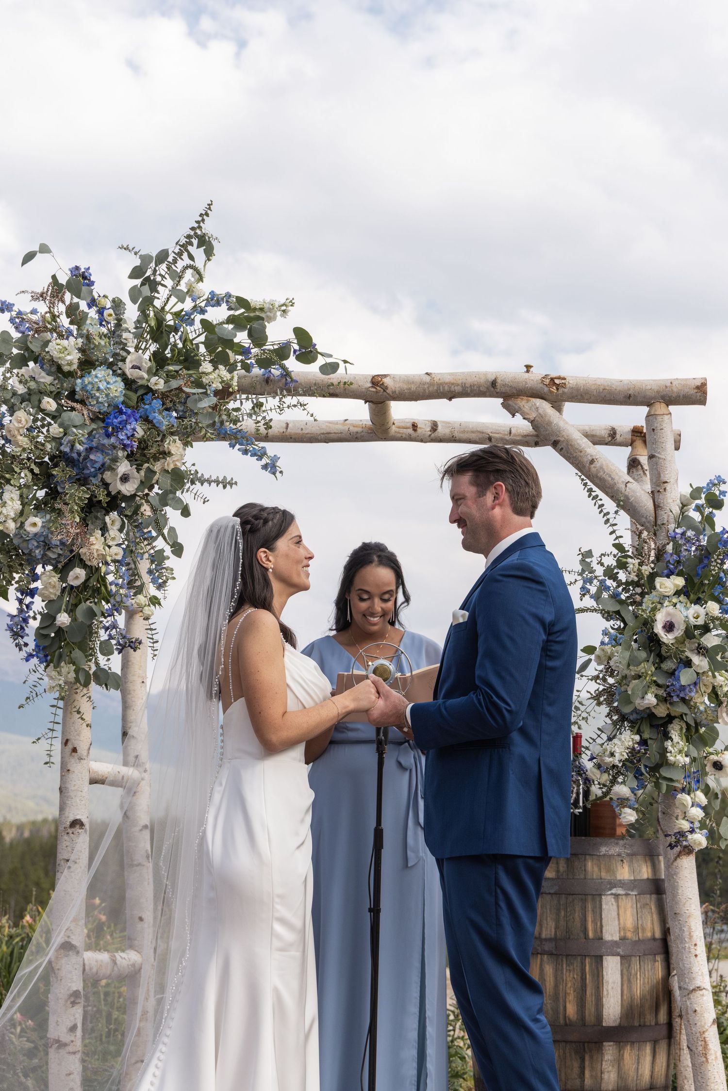 Bride and groom exchange vows under a rustic birch arch adorned with blue and white flowers at an outdoor wedding.