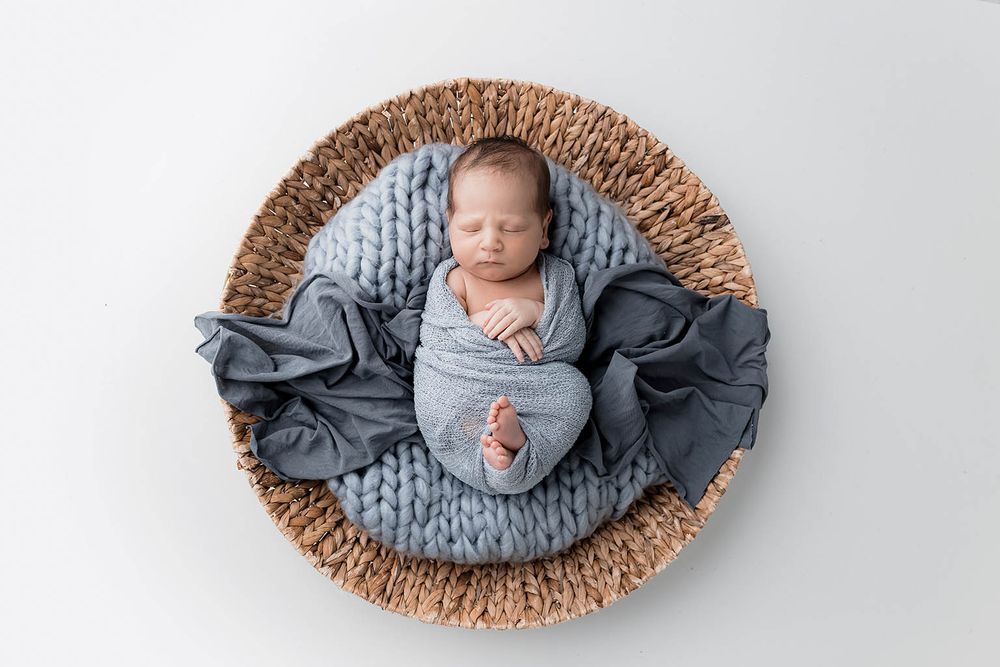 Sleeping newborn baby wrapped in blue swaddle nestled in woven basket with knit blanket.
