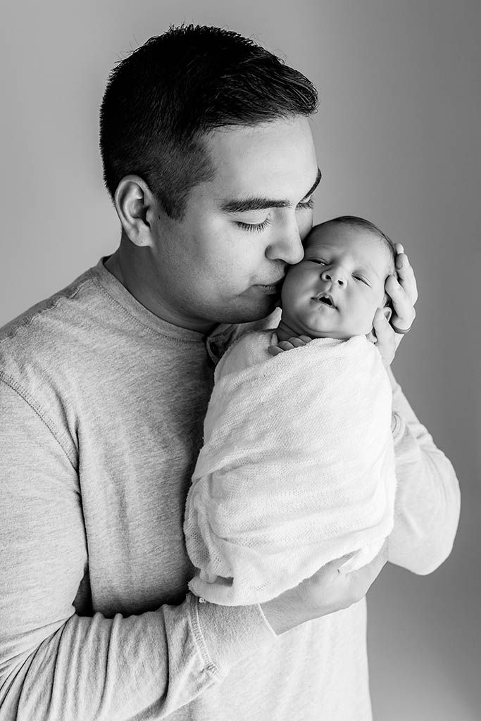 Father gently kissing his swaddled newborn baby in a tender black and white portrait photo.
