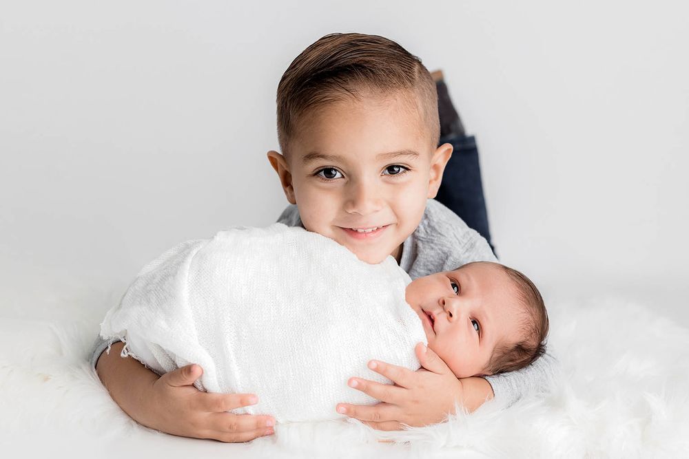 Smiling young boy proudly holds his newborn sibling wrapped in white on a fluffy white surface.