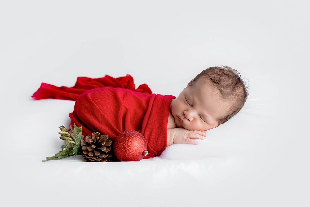 Peaceful newborn baby wrapped in red fabric with Christmas ornament and pinecone decorations.