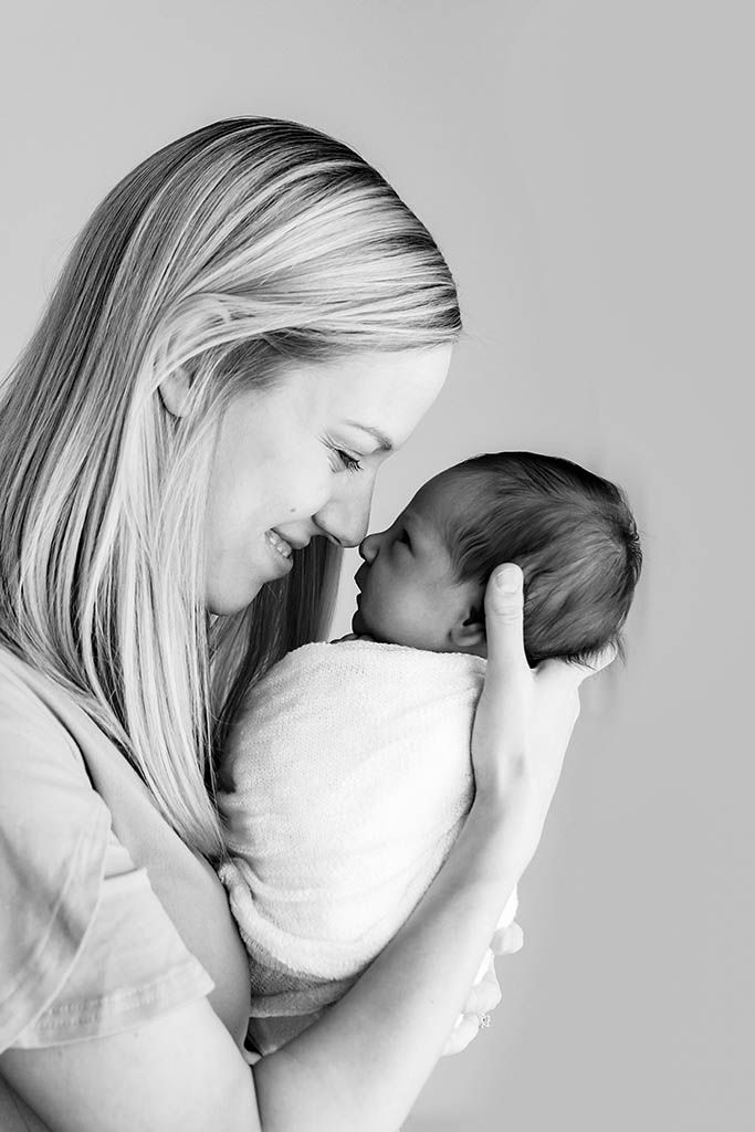 Smiling mother touching noses with her swaddled newborn baby in a black and white portrait.