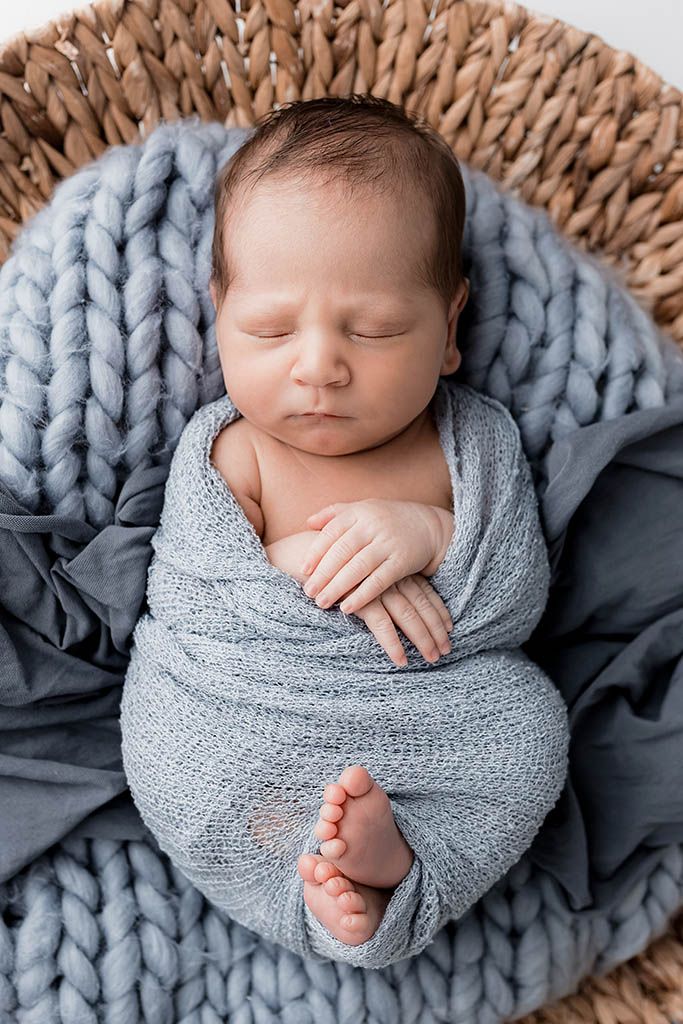 Peaceful newborn baby swaddled in a soft blue wrap, sleeping in a wicker basket with a chunky knit blanket.