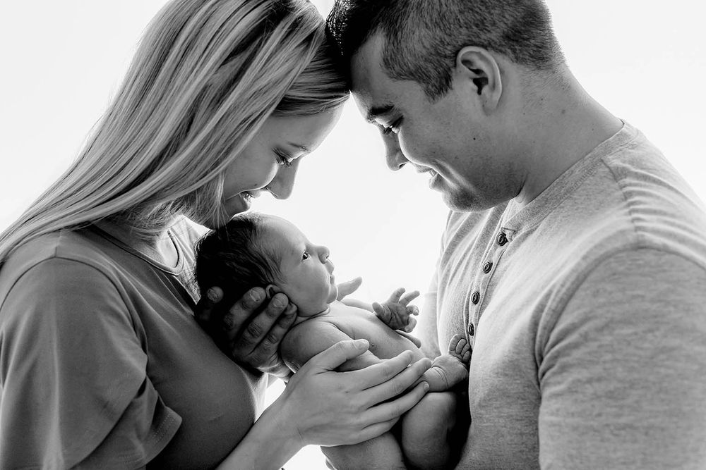 Black and white photo of new parents tenderly holding their newborn baby together, foreheads touching.