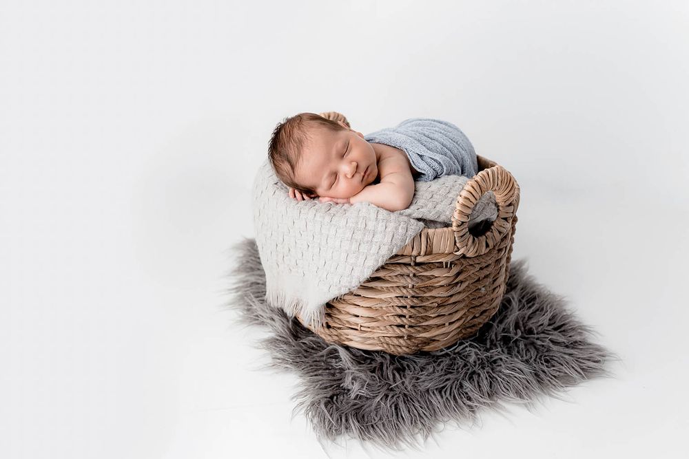 Sleeping newborn baby wrapped in blue swaddle resting in a wicker basket on a gray fur rug.