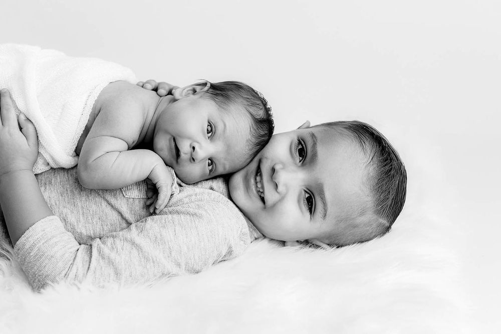 Black and white photo of two babies lying together on a soft white blanket, looking at the camera.