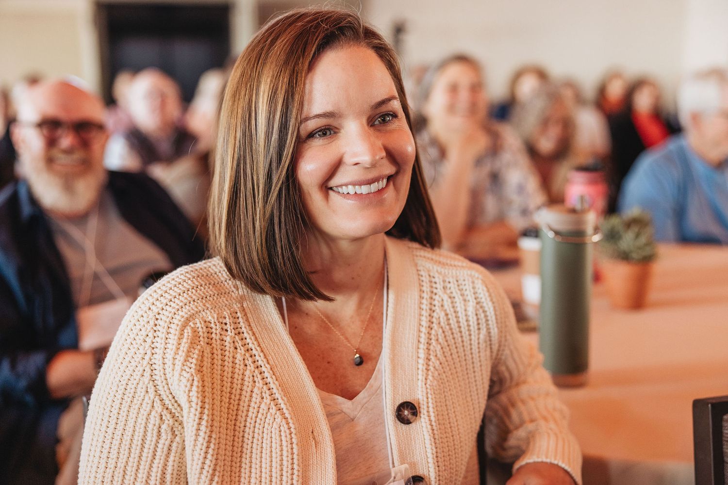 Smiling woman with bob haircut in beige cardigan sitting at a table in a casual gathering setting.