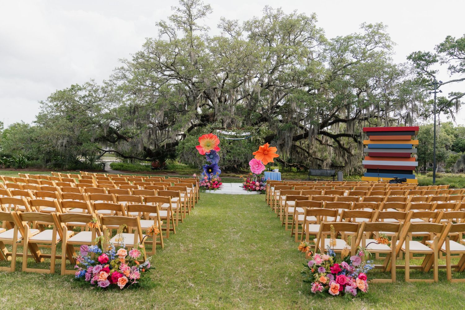 Outdoor wedding ceremony setup with wooden chairs, colorful floral arrangements, and a vibrant arch under a sprawling oak tree.