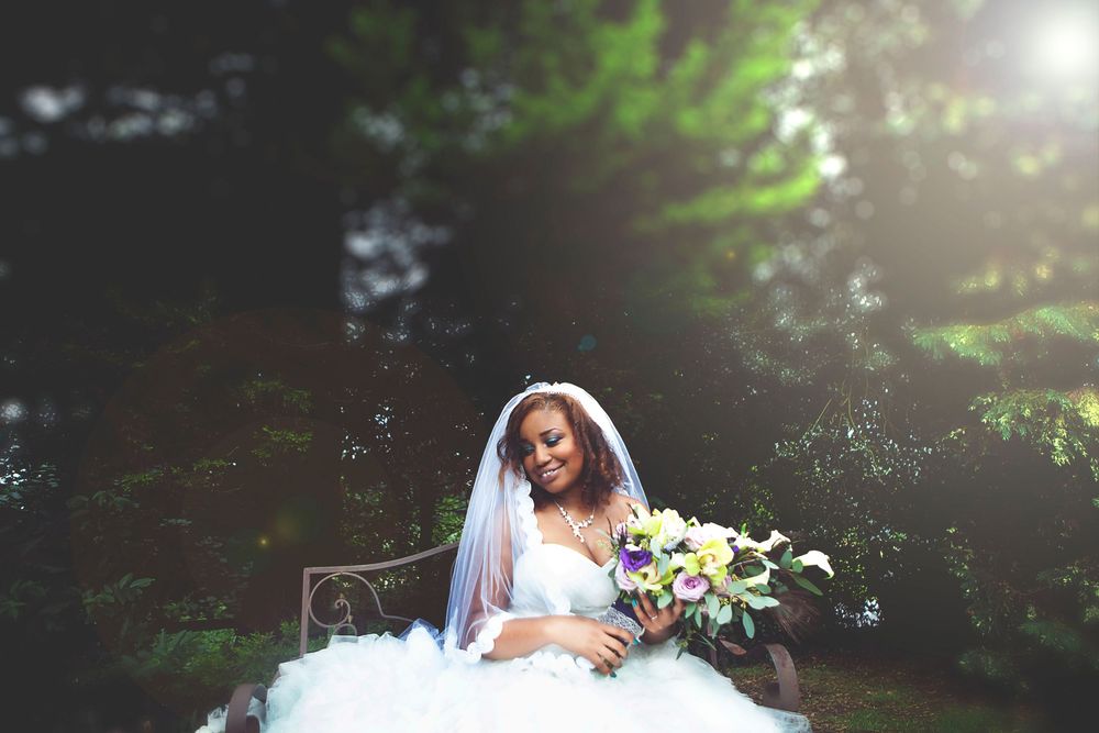 Elegant bride in white ballgown and veil holding a bouquet, seated outdoors surrounded by lush green trees. At Oaks farm wedding venue.