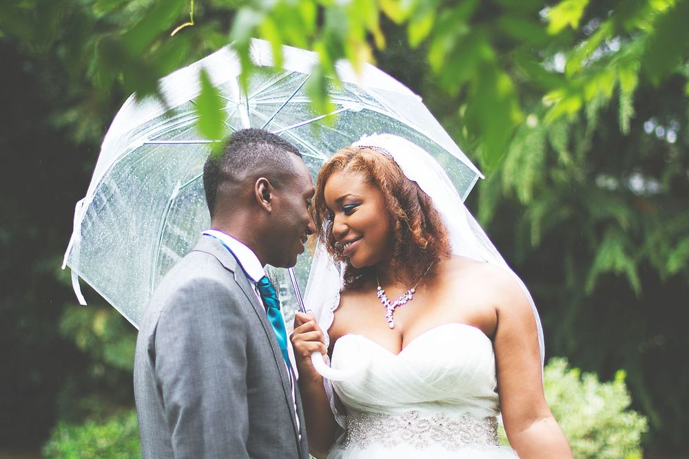 A bride and groom share a romantic moment under a clear umbrella in a lush green garden on their wedding day. At Oaks farm wedding venue.