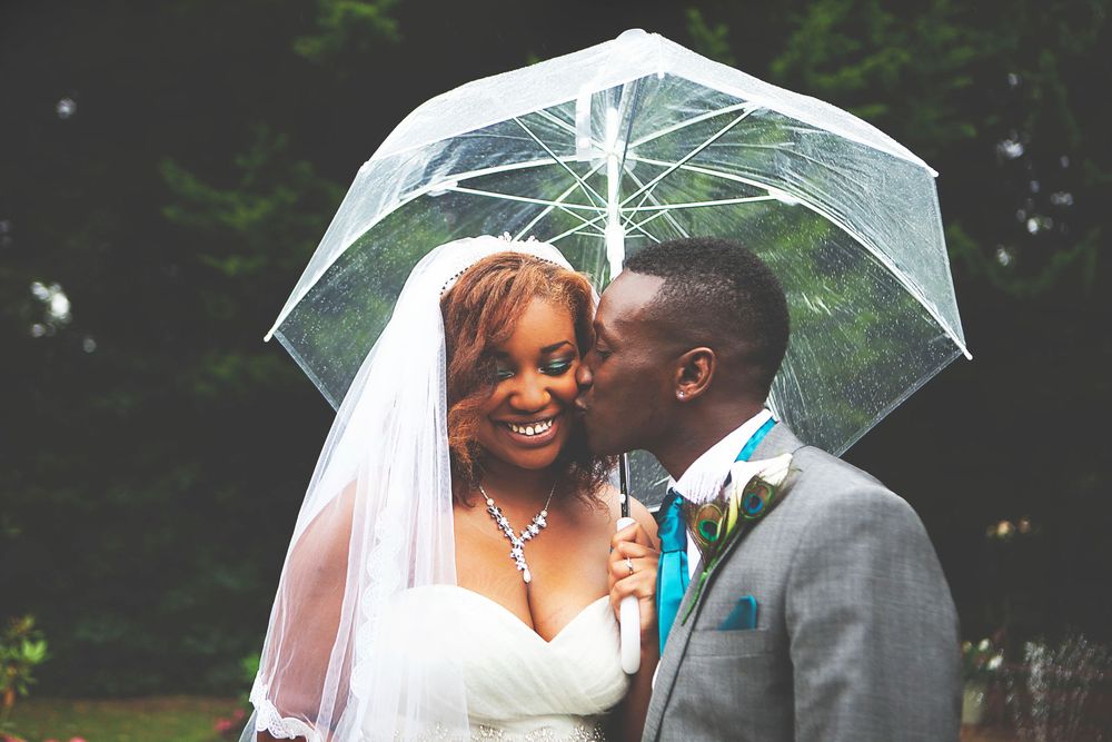 A groom kisses his bride under a clear umbrella in the rain on their wedding day. At Oaks farm wedding venue in Surrey.
