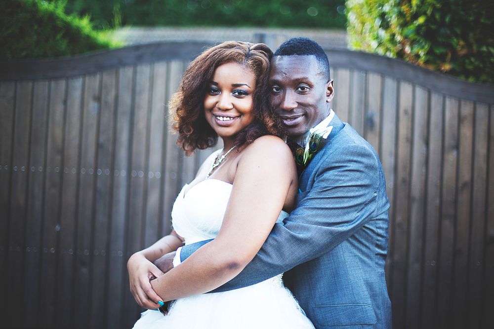 A smiling bride and groom in wedding attire pose together outdoors near a wooden gate. At Oaks farm wedding venue in Surrey.