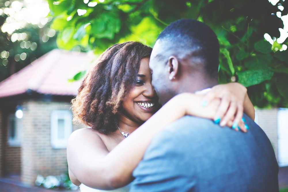 A smiling couple sharing a warm embrace outdoors, with lush green trees and a brick building in the background. At Oaks farm wedding venue.