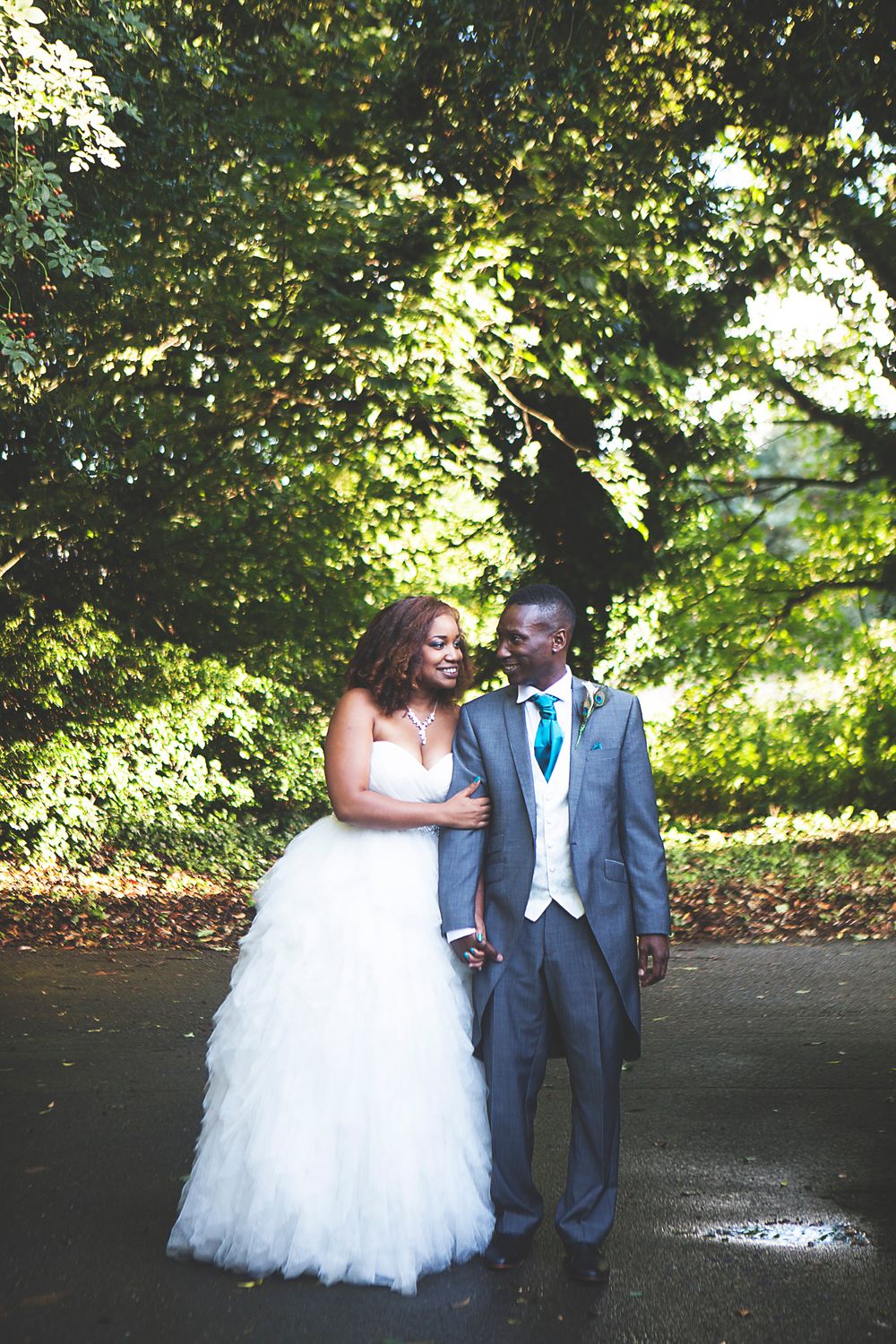 A bride in a white tulle gown and groom in a gray suit share a romantic moment outdoors surrounded by lush green trees. At Oaks farm venue.