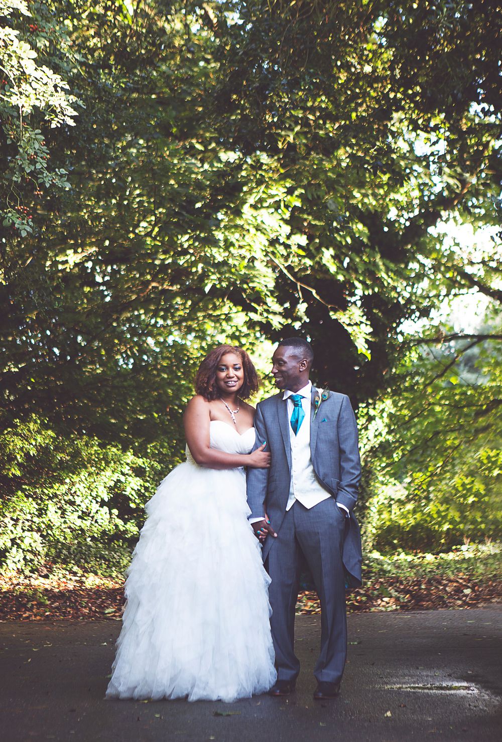 Bride in white gown and groom in gray suit with teal tie pose together outdoors under lush green trees on their wedding day at Oaks farm.