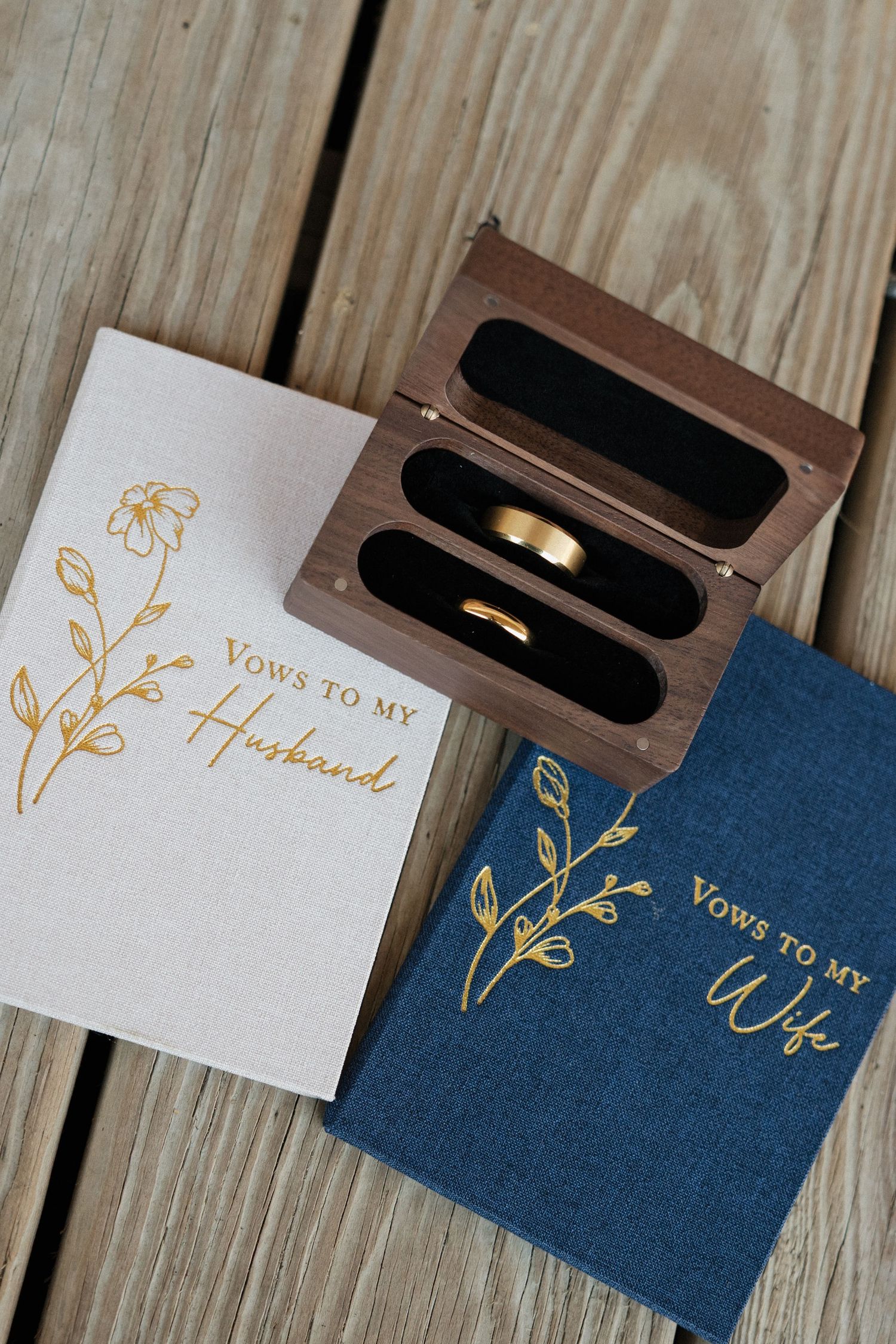 Wedding vow books for husband and wife with gold floral designs beside a wooden ring box holding two gold bands.