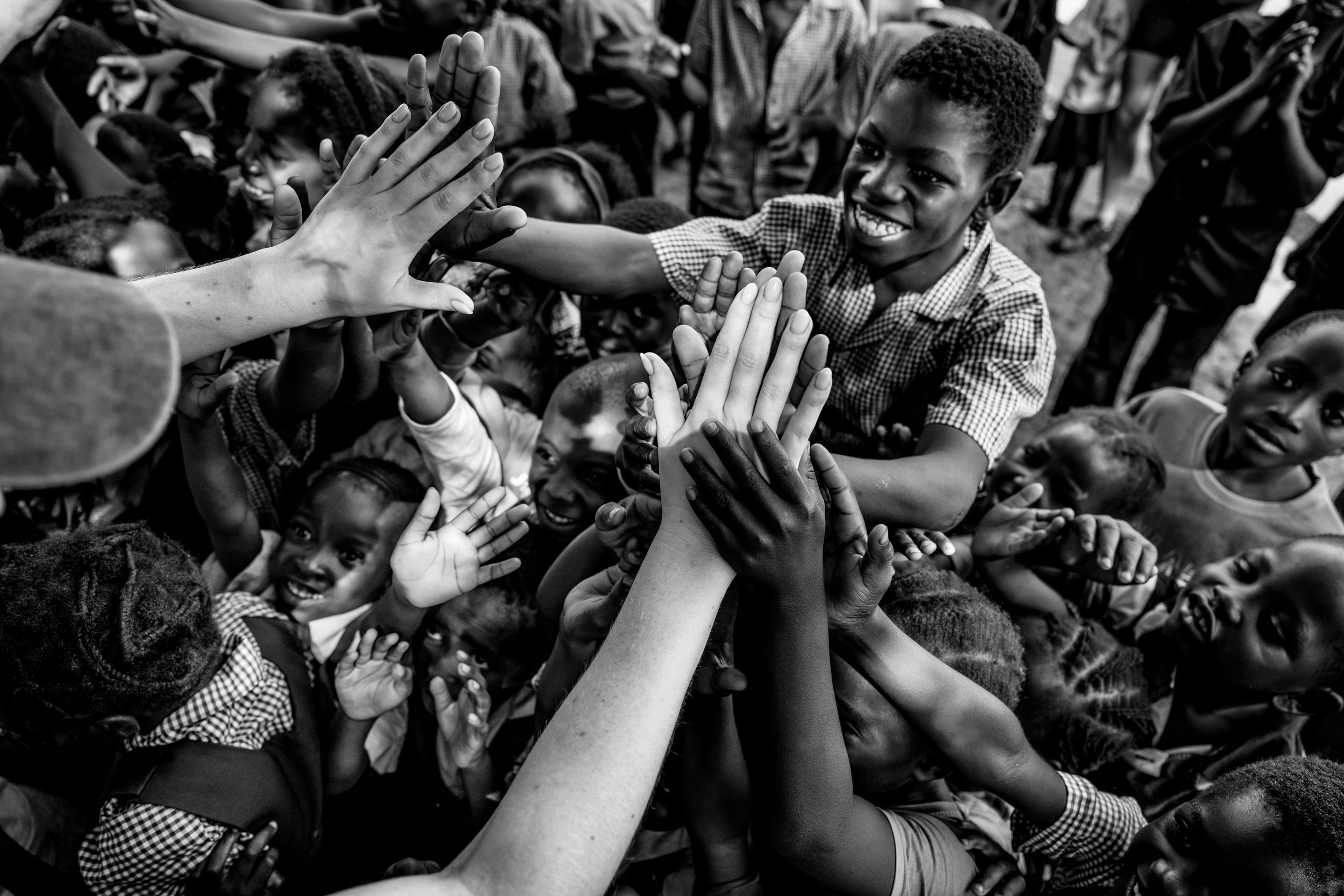 Black and white photo of African children eagerly reaching out their hands toward a visitor in a crowded outdoor gathering.