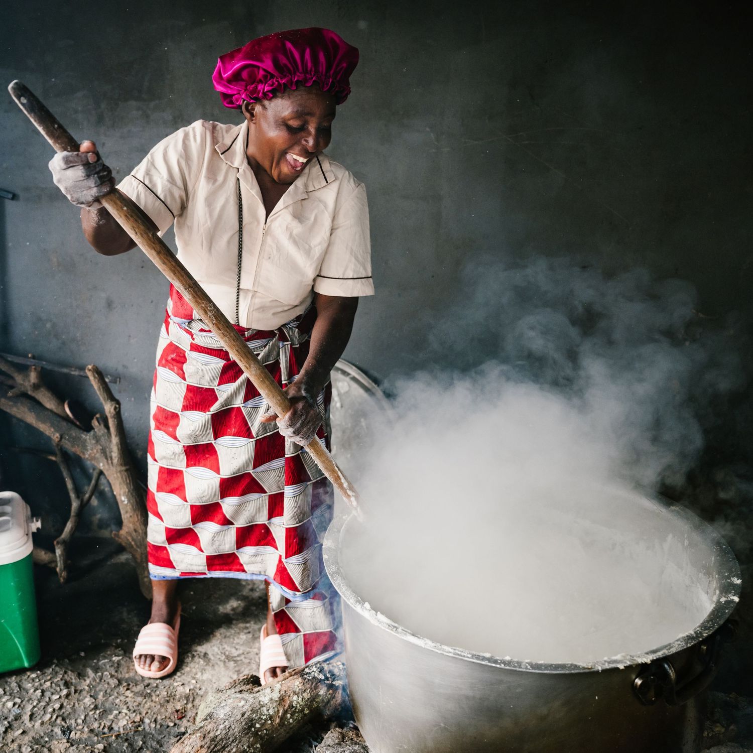 African woman in traditional attire stirring large steaming pot over open fire in rustic outdoor kitchen.