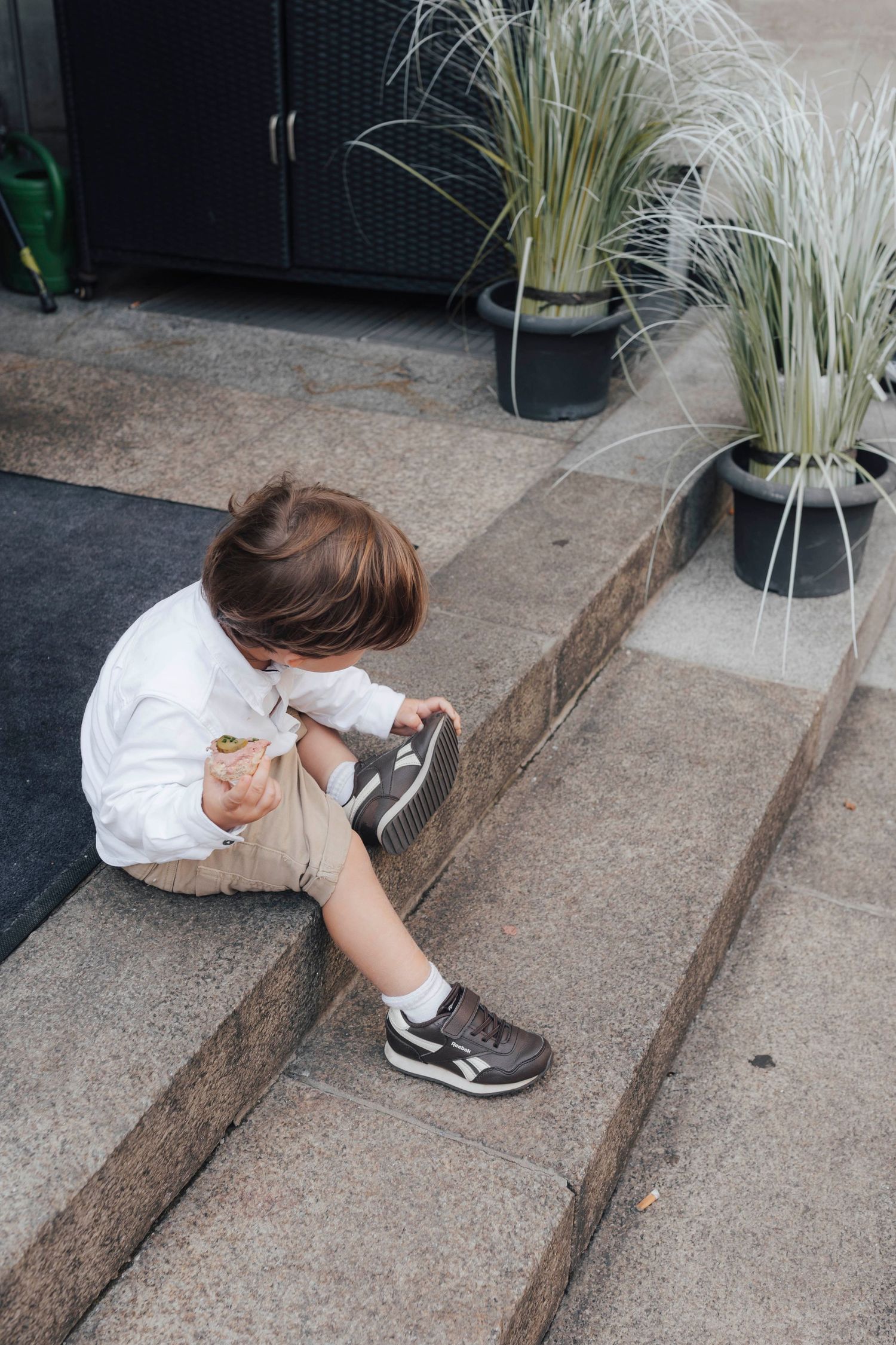 Young toddler boy sitting on outdoor steps, tying his sneakers while holding a snack, with potted plants nearby.