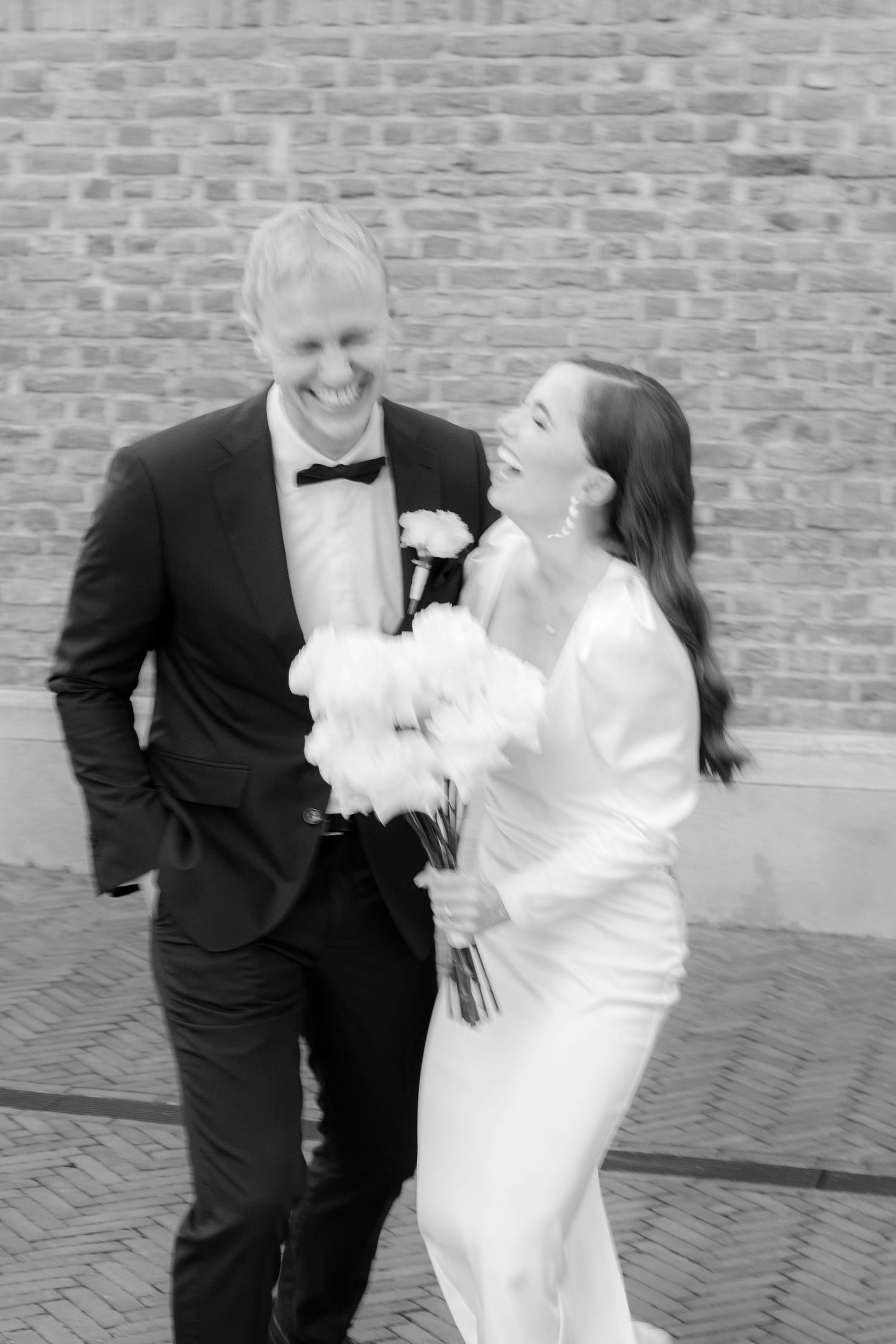 Black and white photo of a laughing bride and groom walking together, she holds white flowers, brick wall background.