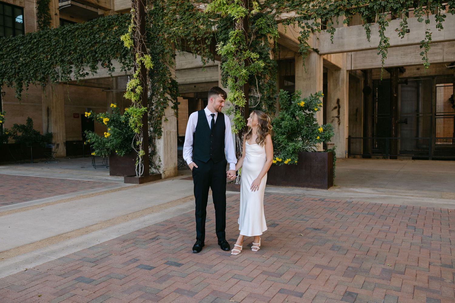 Couple in wedding attire holding hands outside a building with lush green ivy walls on a brick plaza.