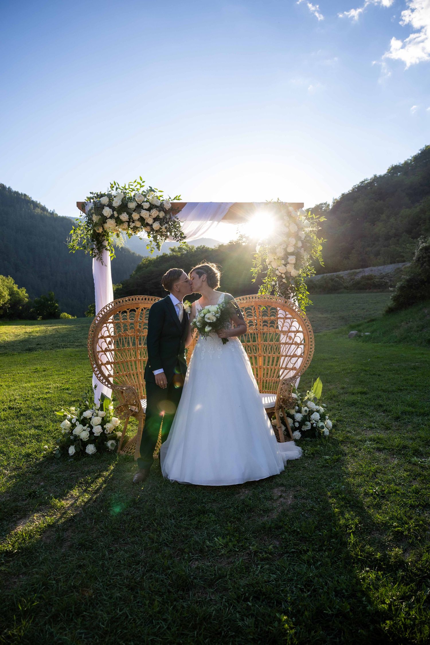 Bride and groom kissing at outdoor wedding altar with floral arch, wicker chairs, and mountain backdrop at sunset.