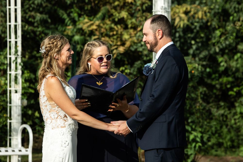 Bride and groom exchange vows outdoors with officiant in sunglasses holding book during sunny wedding ceremony.
