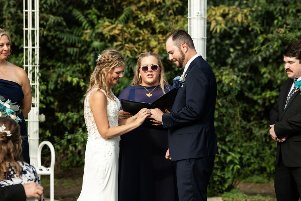 Bride and groom exchange rings during outdoor wedding ceremony with officiant under white arbor surrounded by greenery.