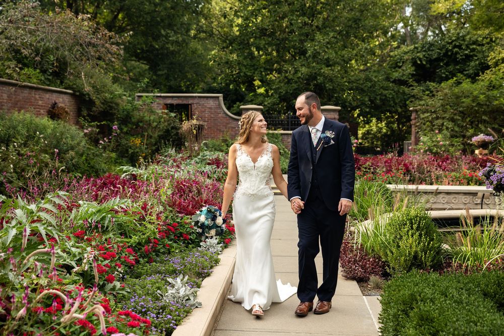 Bride and groom walking hand in hand through a lush garden path surrounded by colorful flowers on their wedding day.