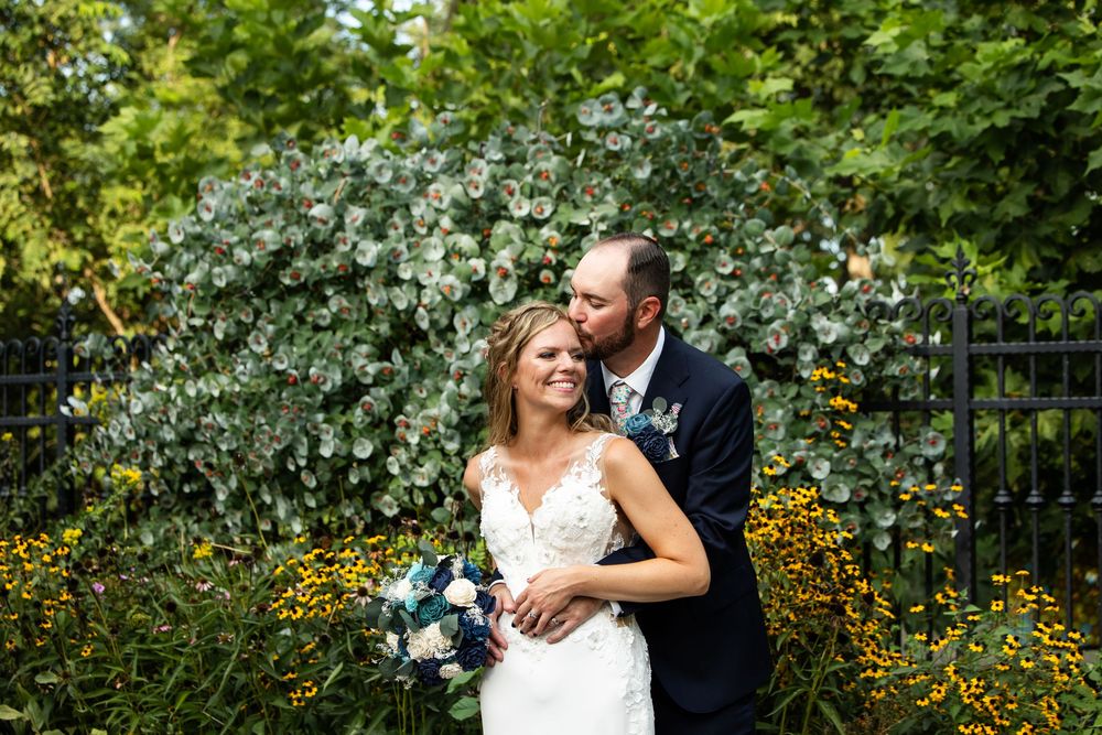 Groom kisses bride on cheek as she holds bouquet in front of lush green garden with yellow flowers.