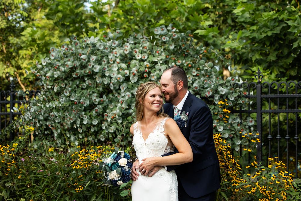 Bride and groom embracing outdoors, groom kissing bride's cheek as she smiles holding a blue and white bouquet.