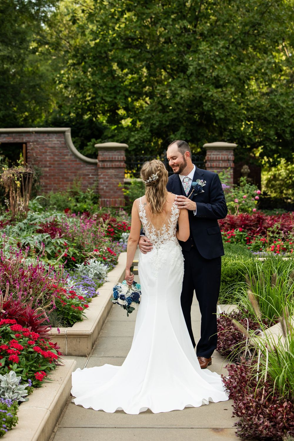 Bride and groom walk through a colorful garden, she in a lace gown holding bouquet, he in a navy suit.