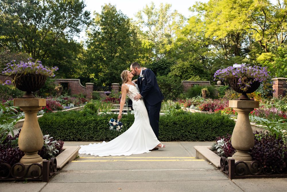 Bride and groom sharing a romantic kiss in a lush garden wedding venue with purple flowers and manicured hedges.