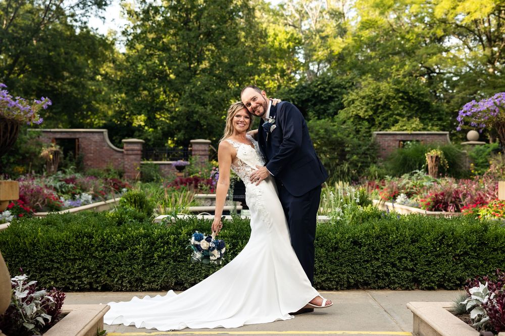 Bride and groom pose together in a lush garden wedding portrait, bride holding blue bouquet in white gown.