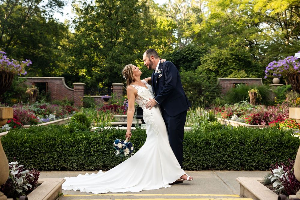 Bride and groom share a romantic kiss in a lush garden setting, bride holding a blue bouquet in a white gown.