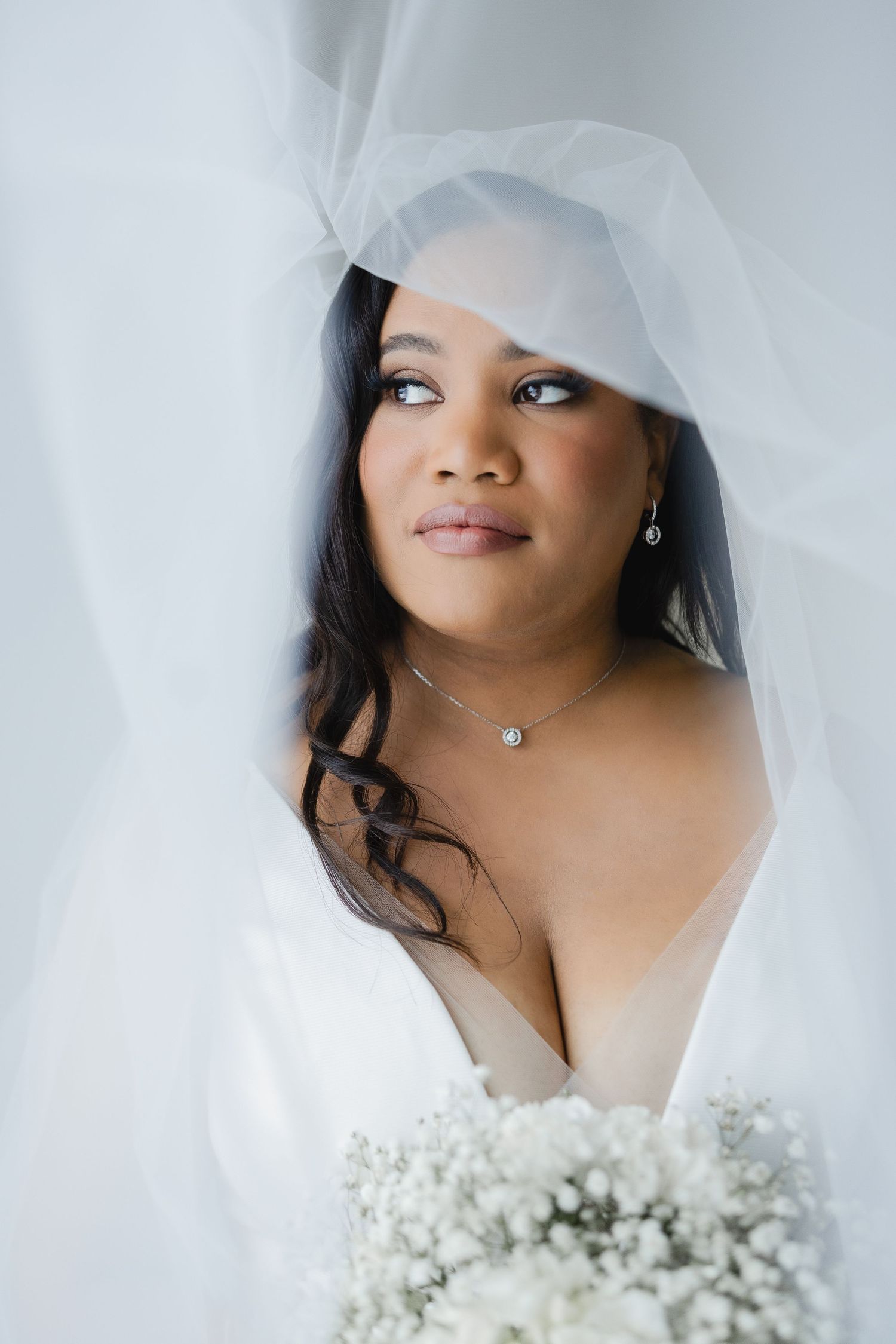 Bride in white veil and gown holds baby's breath bouquet, wearing diamond necklace and earrings.