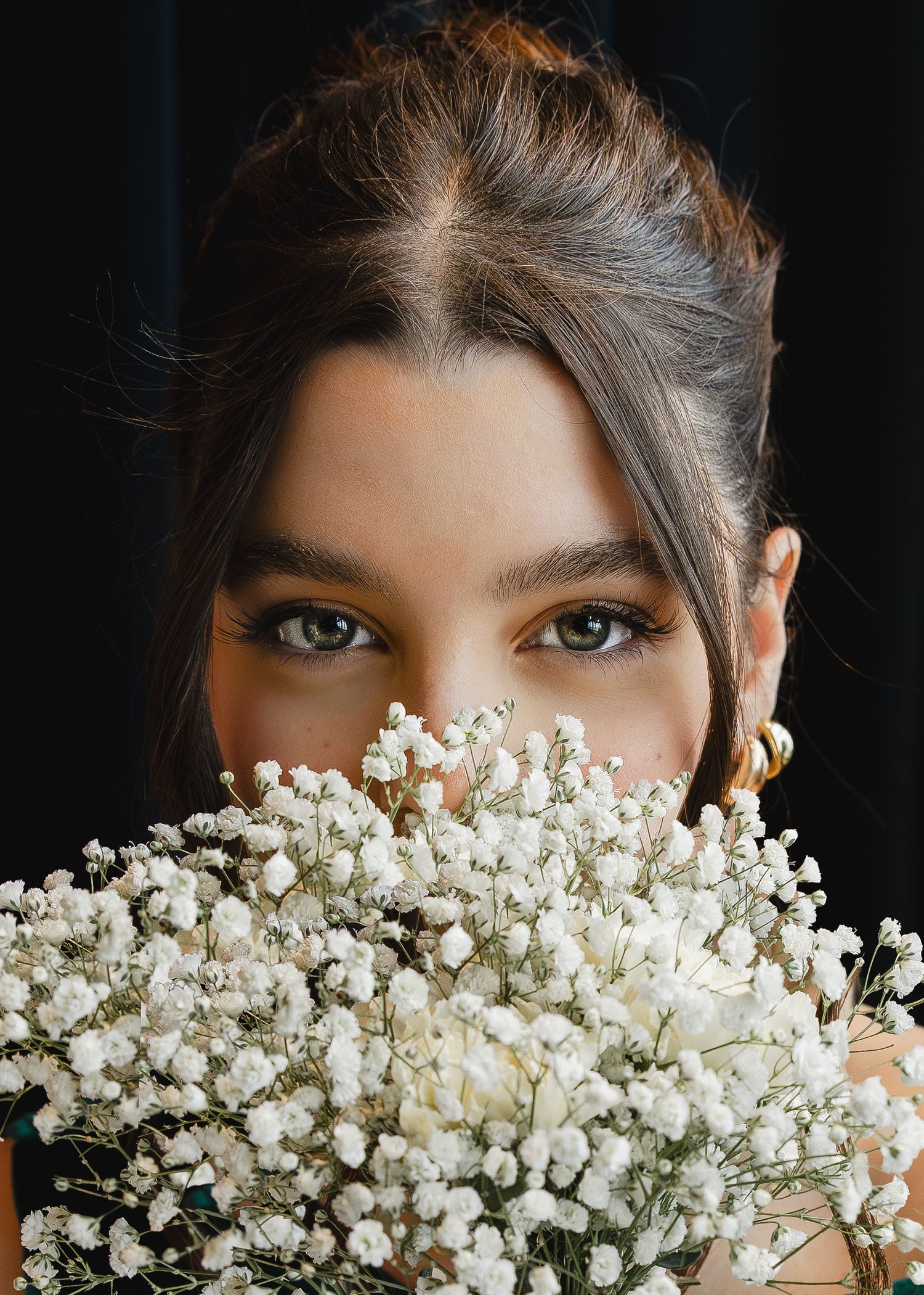 Young woman with updo hairstyle holding white baby's breath flowers covering her face, wearing gold earrings.