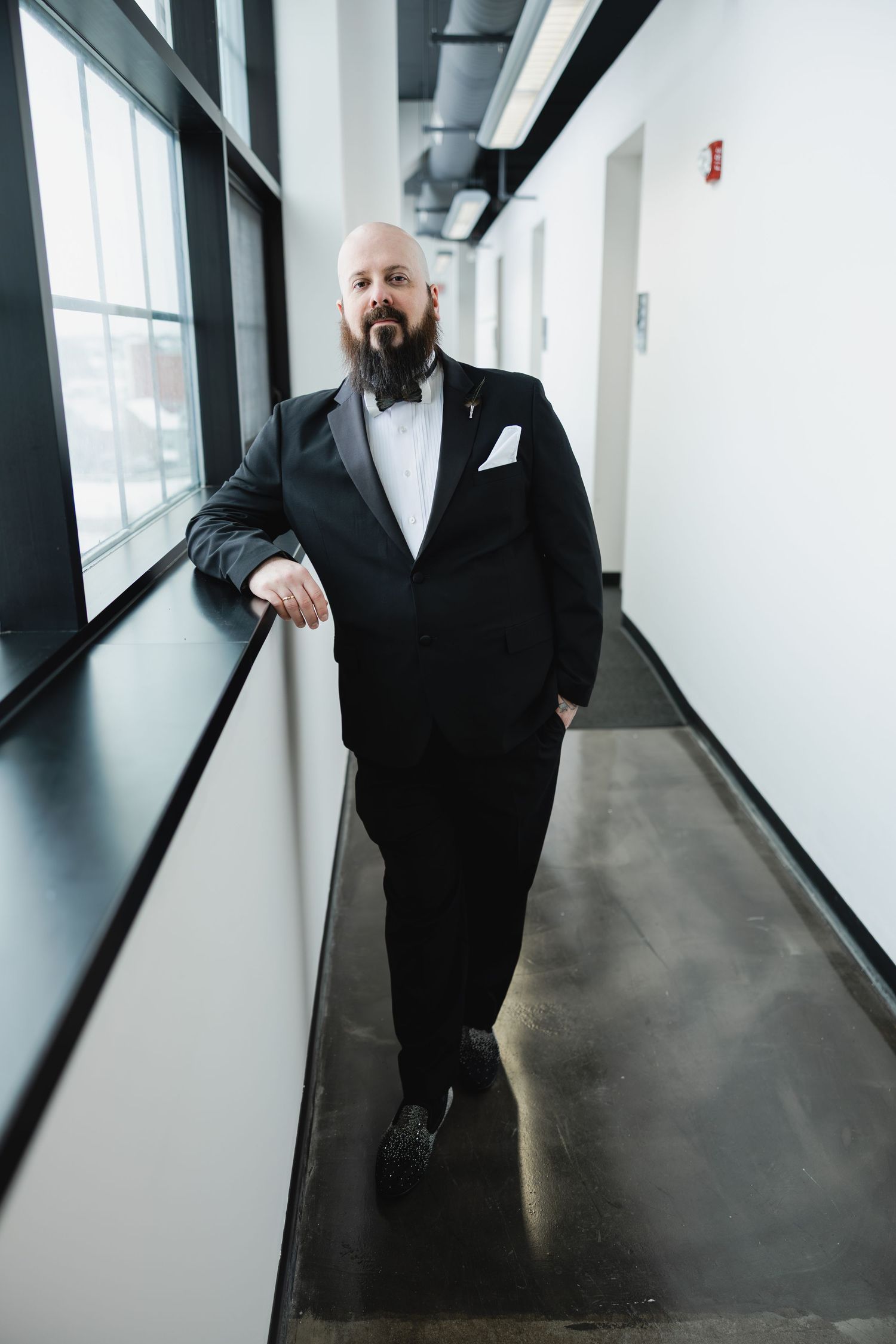Bald groom with beard in black tuxedo and bow tie leans against wall in modern corridor.