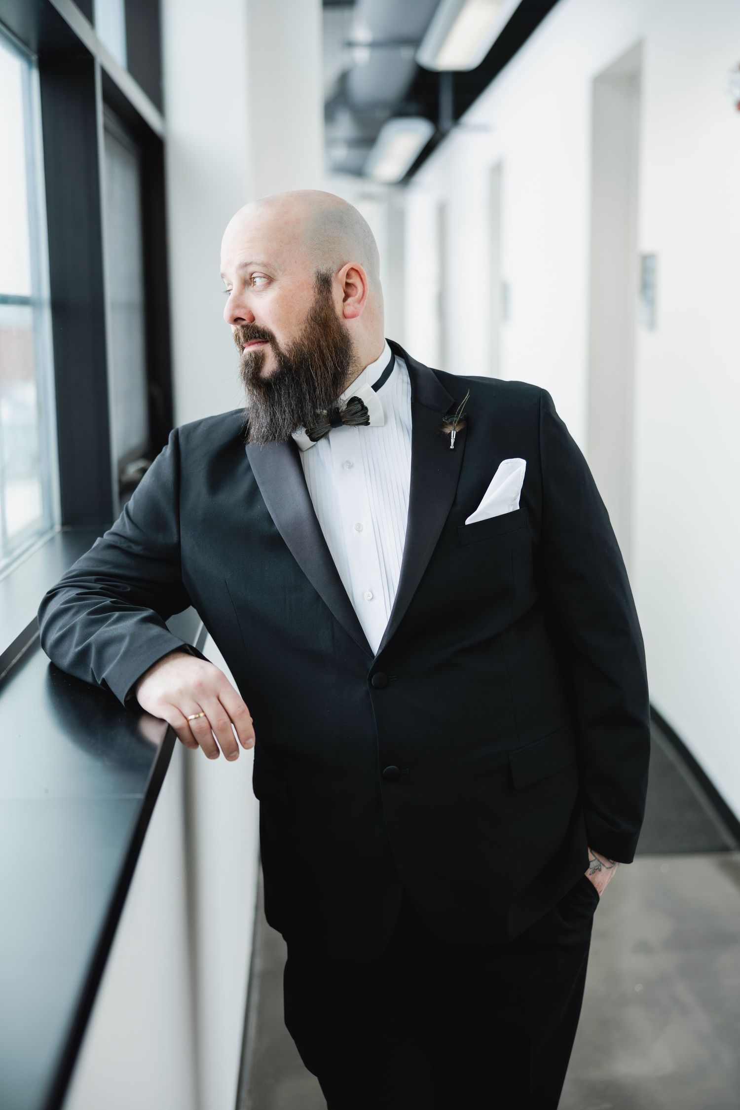 Bald bearded groom in black tuxedo with boutonniere gazes out window in elegant hallway.