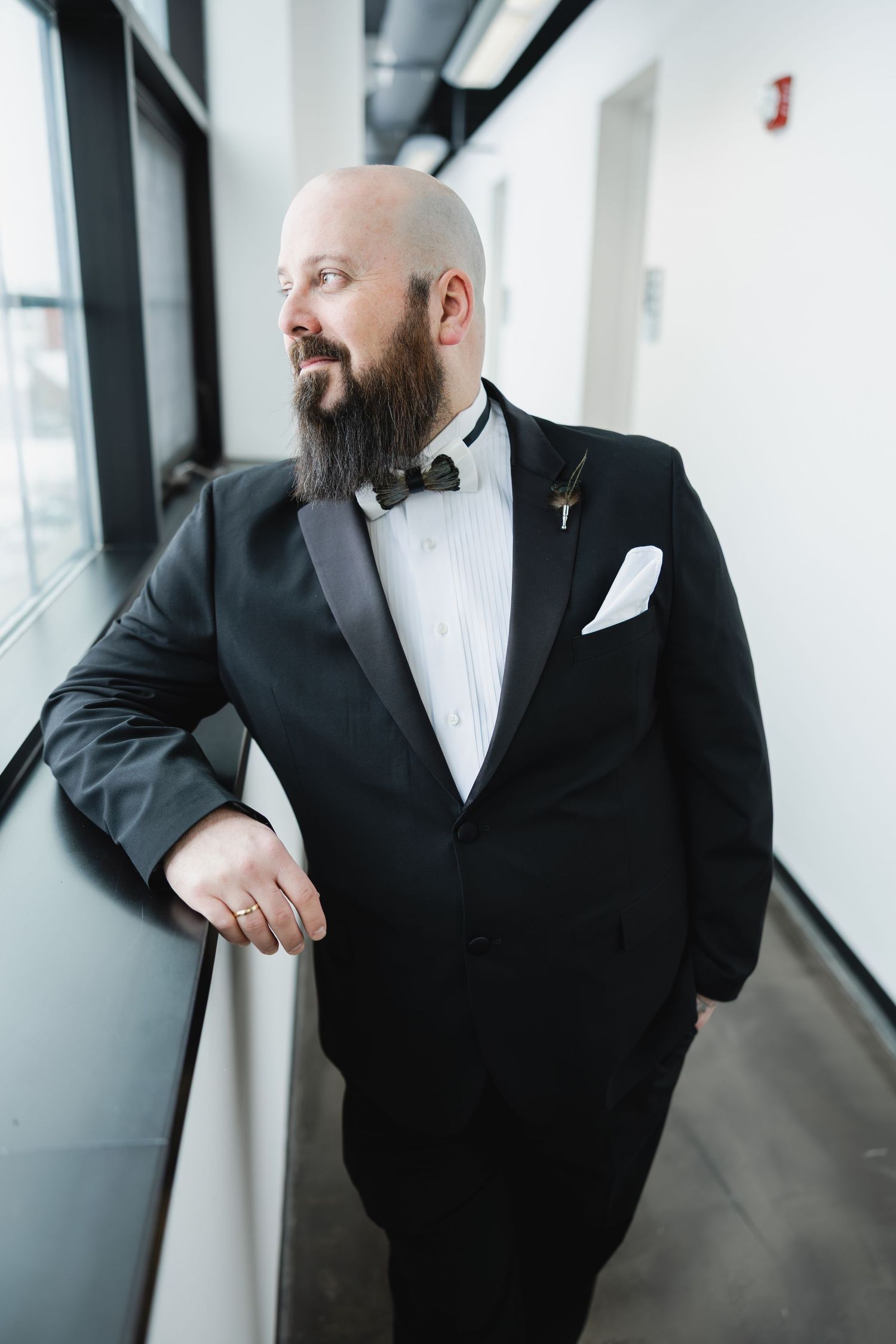 Bald groom with beard in black tuxedo and bow tie leans against railing near window on wedding day.