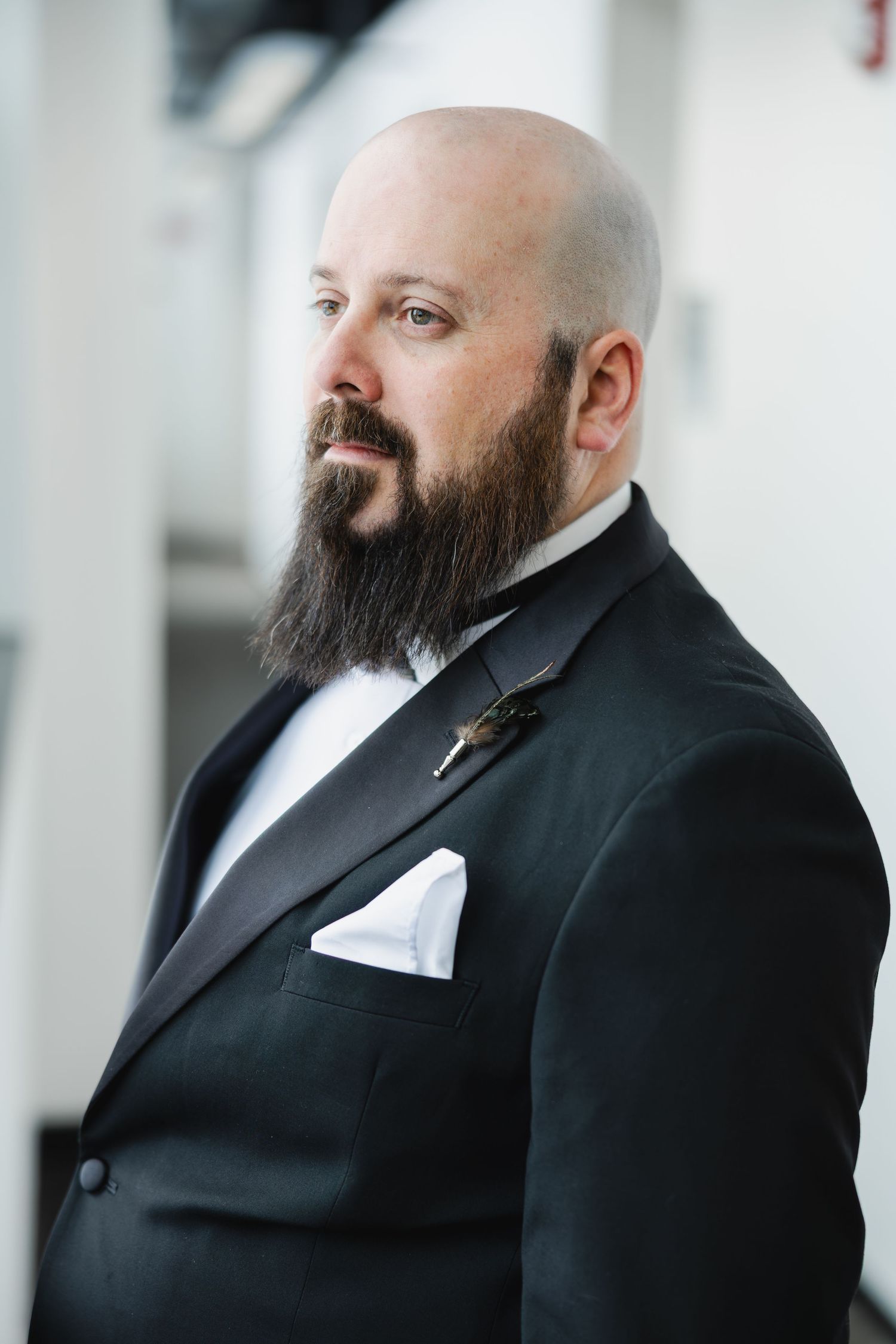 Bald groom with a long beard poses in a classic black tuxedo with white pocket square and boutonniere.