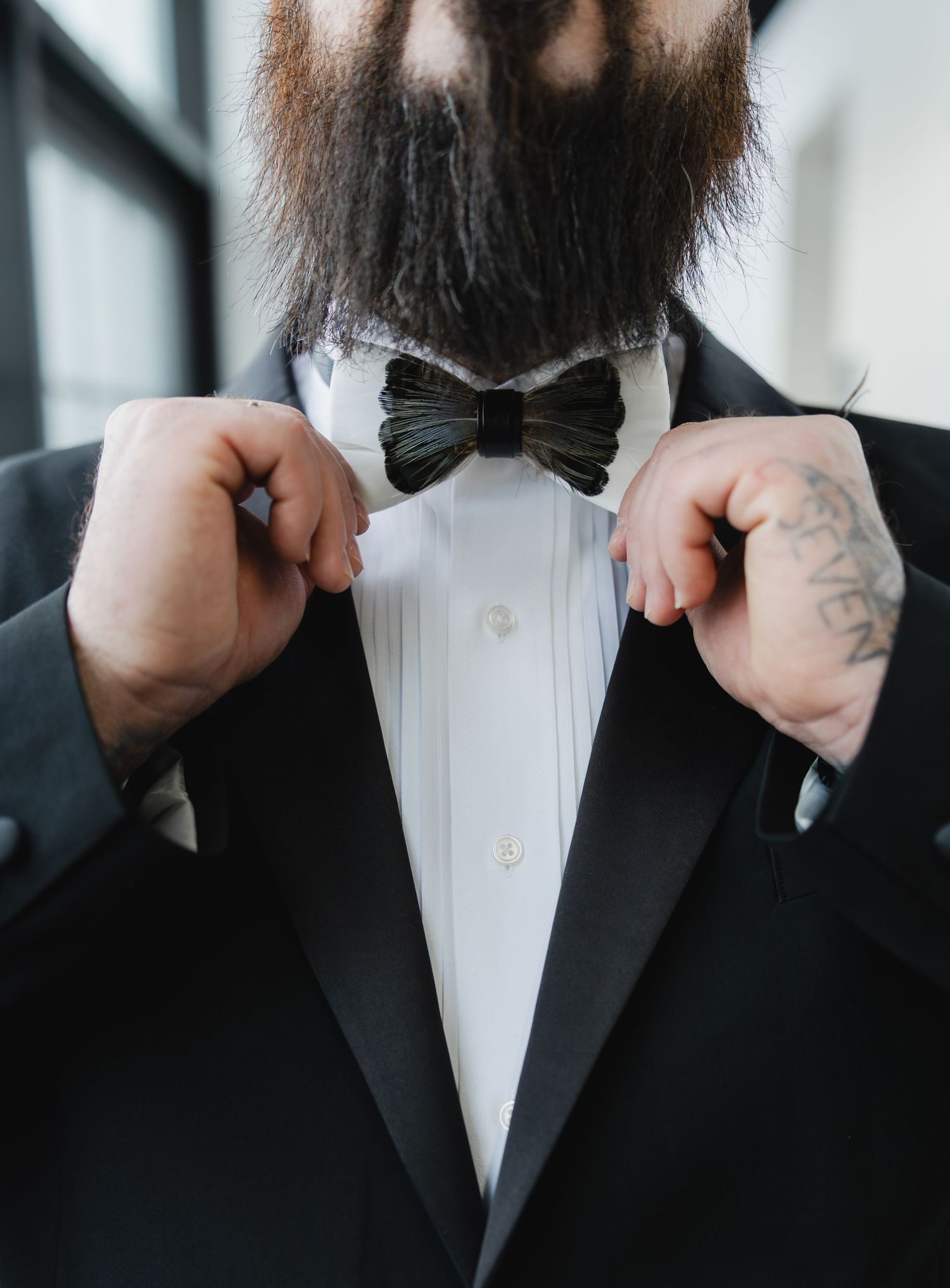 Bearded man in black tuxedo adjusting bow tie, tattoo visible on hand, formal wedding attire.
