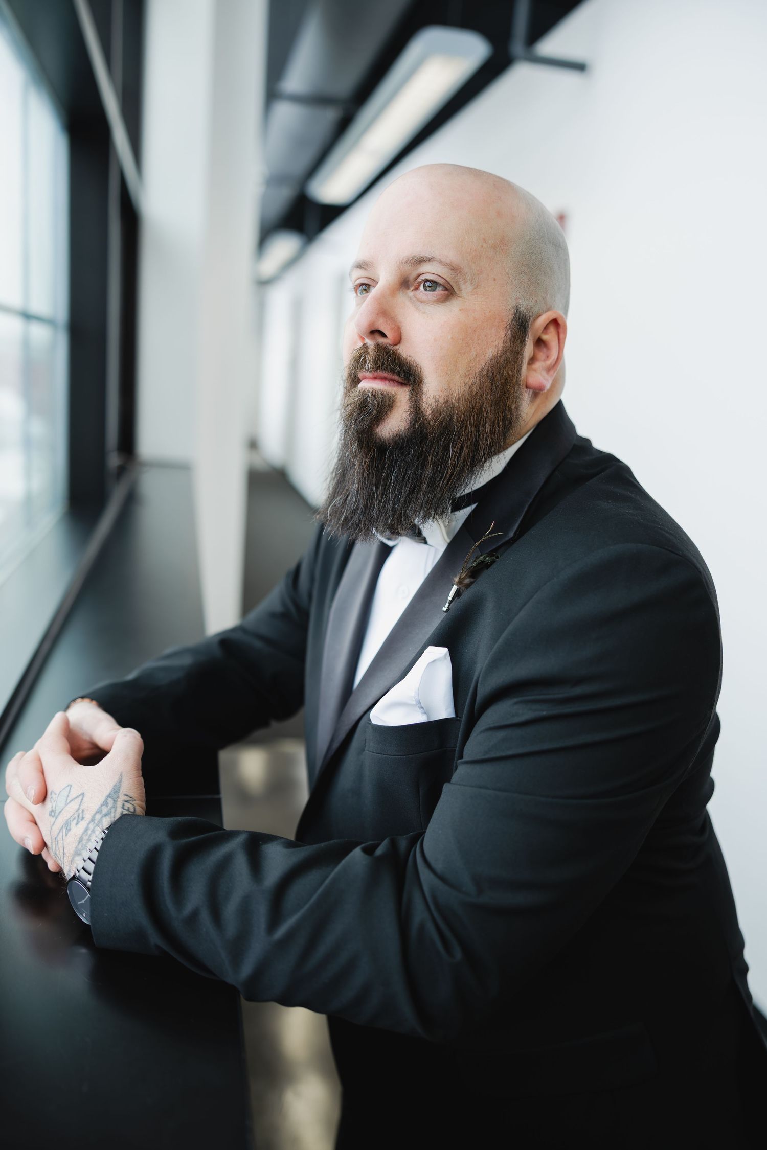 Bald groom with long beard in black tuxedo and bow tie poses thoughtfully by window.