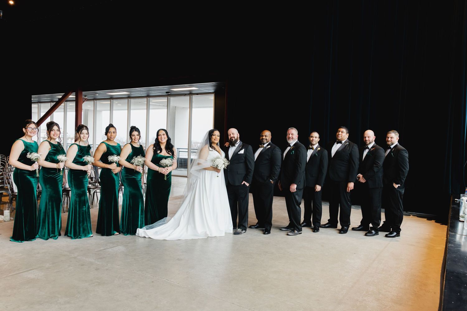 Wedding party poses on stage, bridesmaids in emerald green velvet gowns beside bride in white and groomsmen in black tuxedos.